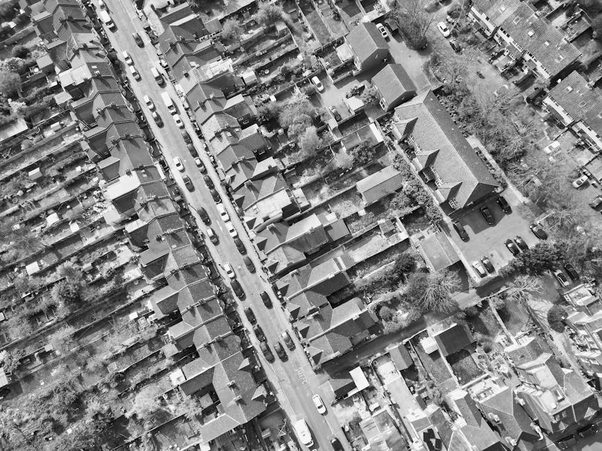 Aerial black-and-white photograph of a residential street lined with terraced houses and detached homes, showing driveways, parked cars, trees, and rooftops with varying roof styles. The image capture