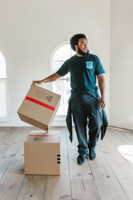 Close-up of a person wearing a dark blue t-shirt and dark trousers, securing a cardboard box with a caution label that reads 'CAUTION THIS SIDE UP' with a red arrow pointing upward. The individual is