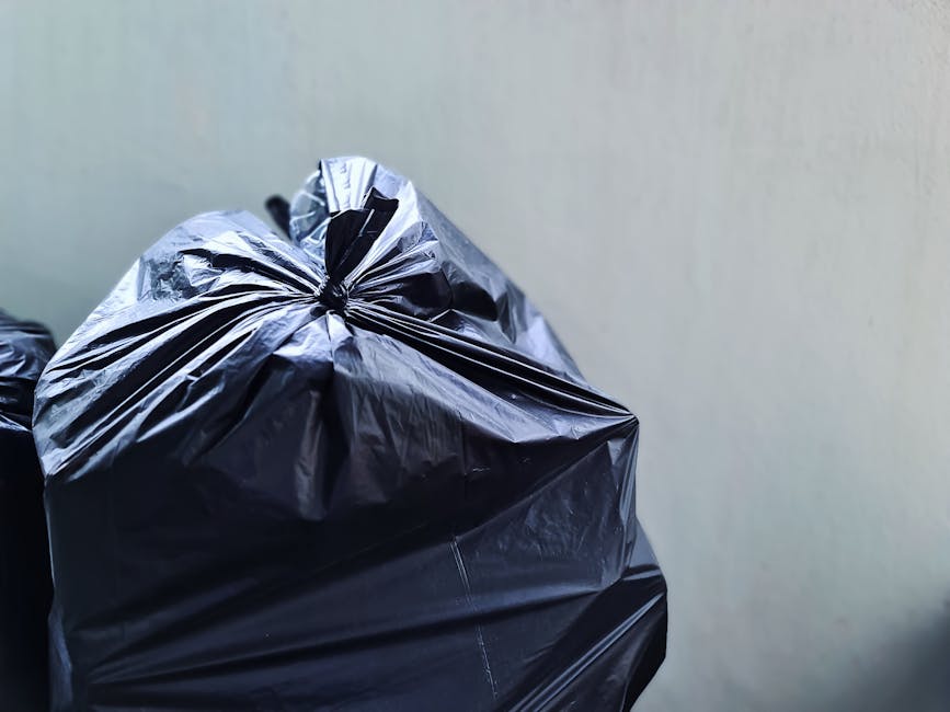 A person wearing a white shirt and beige trousers is seen placing a clear plastic bottle into a grey recycling bin labeled 'PLASTIC' in a domestic setting. The person has a red-orange knitted scarf dr