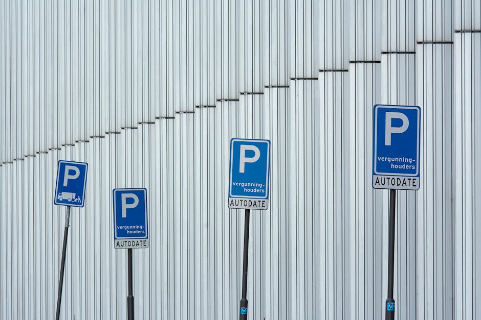 Close-up view of a yellow rectangular sign mounted on a metal wire fence, displaying a red and blue no-parking symbol and the German text 'Parken verboten!' which translates to 'No parking!'. Below th